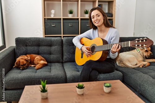 Young hispanic woman playing classical guitar sitting on sofa with dogs at home