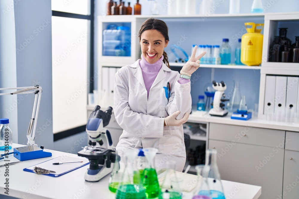 Young brunette woman working at scientist laboratory smiling with happy face winking at the camera doing victory sign. number two. Young brunette woman working at scientist laboratory smiling with happy face winking at the camera doing victory sign. number two.