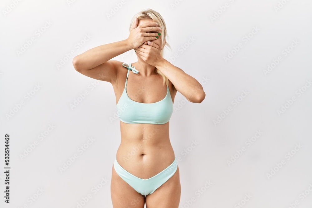 Young caucasian woman wearing bikini over isolated background covering eyes and mouth with hands, surprised and shocked. hiding emotion Young caucasian woman wearing bikini over isolated background covering eyes and mouth with hands, surprised and shocked. hiding emotion