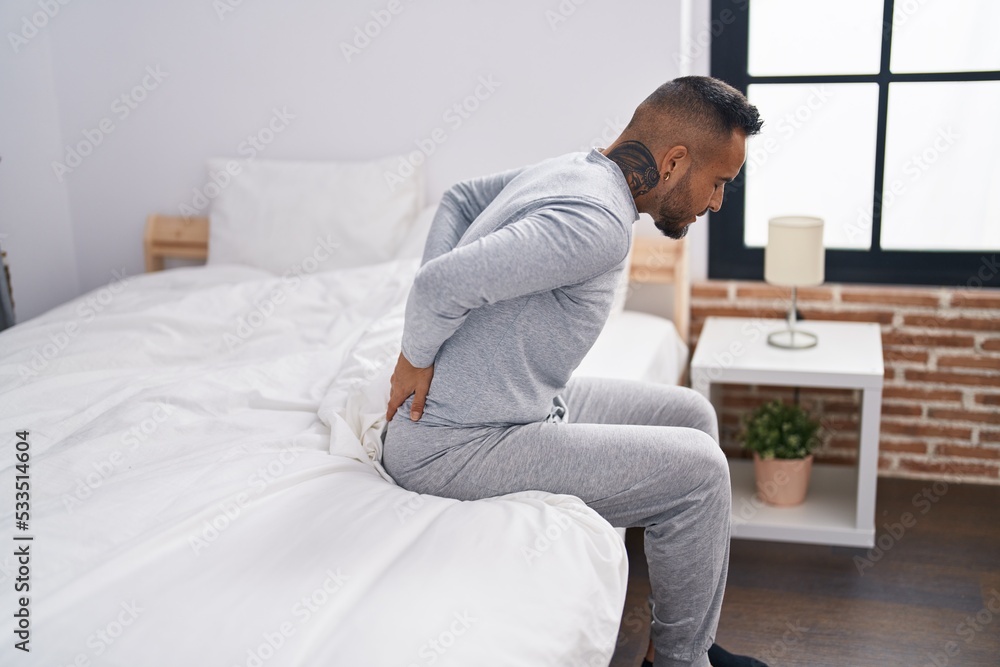 Young hispanic man suffering for backache sitting on bed at bedroom Young hispanic man suffering for backache sitting on bed at bedroom