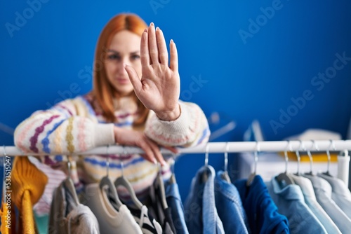 Young woman searching clothes on clothing rack with open hand doing stop sign with serious and confident expression, defense gesture