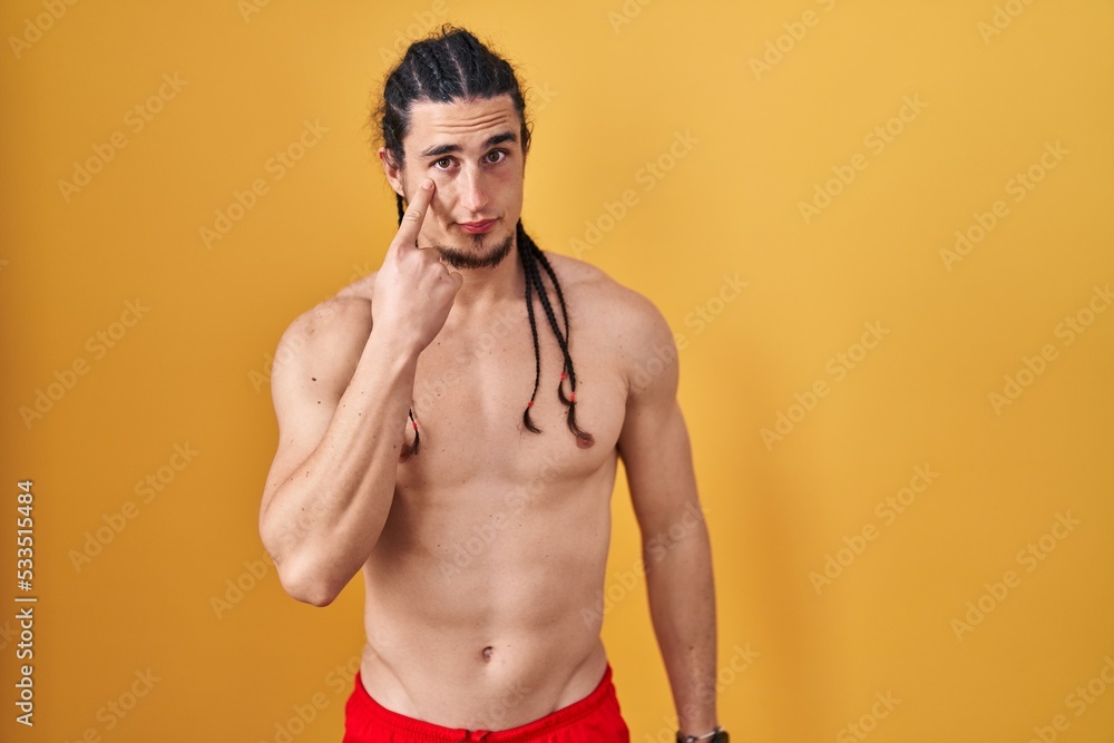 Hispanic man with long hair standing shirtless over yellow background pointing to the eye watching you gesture, suspicious expression