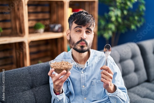 Young hispanic man with beard eating healthy whole grain cereals looking at the camera blowing a kiss being lovely and sexy. love expression.
