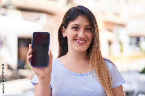 Young hispanic woman smiling confident showing smartphone screen at street