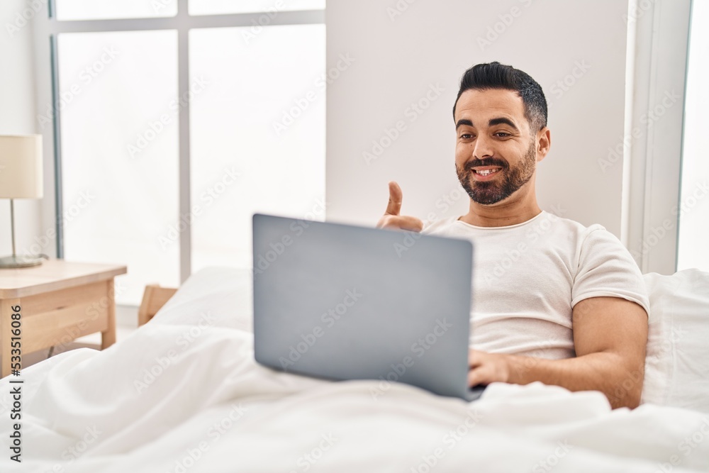 Young hispanic man with beard using computer laptop on the bed smiling happy and positive, thumb up doing excellent and approval sign