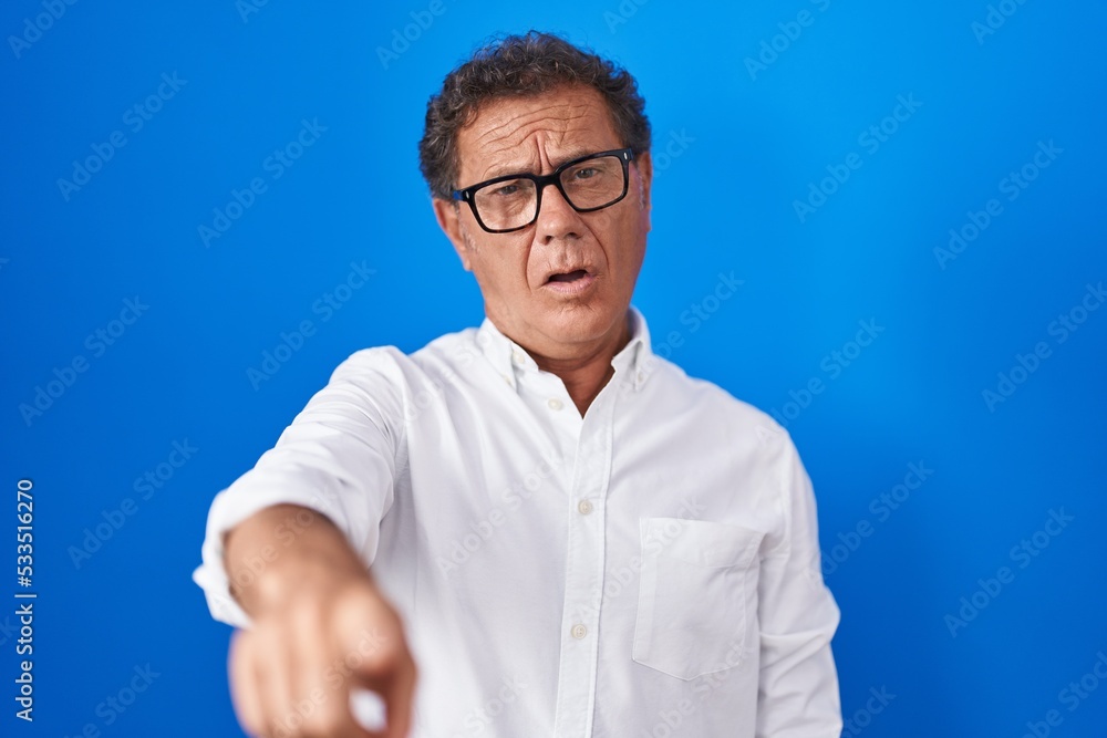 Middle age hispanic man standing over blue background pointing displeased and frustrated to the camera, angry and furious with you Middle age hispanic man standing over blue background pointing displeased and frustrated to the camera, angry and furious with you