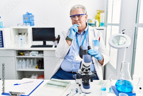 Senior caucasian man working at scientist laboratory looking confident at the camera smiling with crossed arms and hand raised on chin. thinking positive.