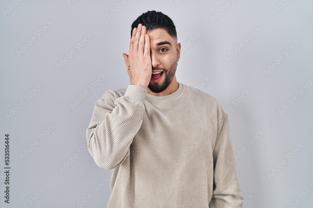 Young handsome man standing over isolated background covering one eye with hand, confident smile on face and surprise emotion. Young handsome man standing over isolated background covering one eye with hand, confident smile on face and surprise emotion.