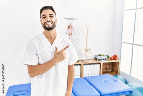 Young handsome man with beard working at pain recovery clinic cheerful with a smile of face pointing with hand and finger up to the side with happy and natural expression on face