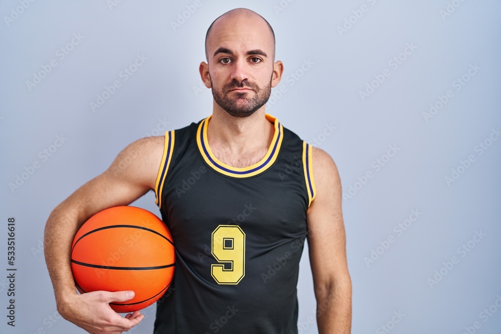 Young bald man with beard wearing basketball uniform holding ball relaxed with serious expression on face. simple and natural looking at the camera.
