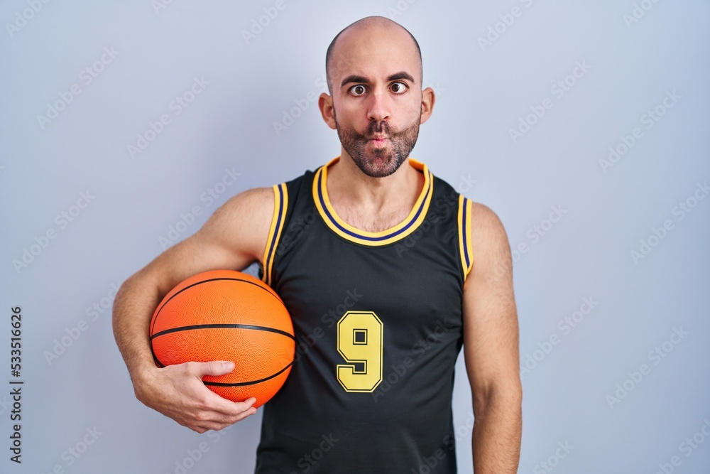 Young bald man with beard wearing basketball uniform holding ball making fish face with lips, crazy and comical gesture. funny expression. Young bald man with beard wearing basketball uniform holding ball making fish face with lips, crazy and comical gesture. funny expression.