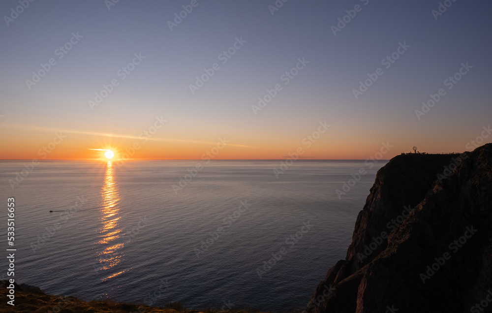 Wonderful landscapes in Norway. Nord-Norge. Beautiful scenery of a midnight sun sunset at Nordkapp (Cape North). Boat and globe on a cliff. Rippled sea and clear orange sky. Selective focus