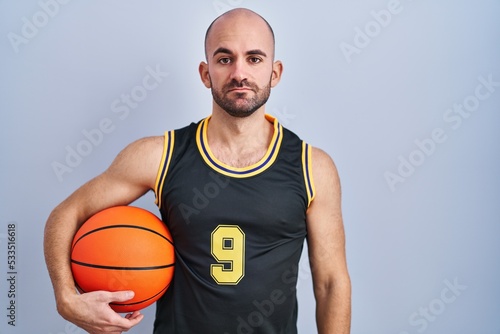 Young bald man with beard wearing basketball uniform holding ball relaxed with serious expression on face. simple and natural looking at the camera.