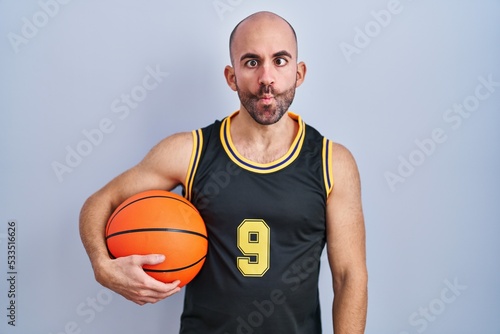 Young bald man with beard wearing basketball uniform holding ball making fish face with lips, crazy and comical gesture. funny expression. © Krakenimages.com
