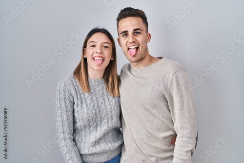 Young hispanic couple standing over white background sticking tongue out happy with funny expression. emotion concept.
