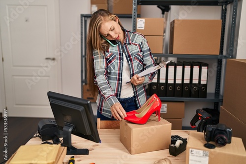 Young blonde woman ecommerce business worker talking on smartphone holding high heel shoes at office