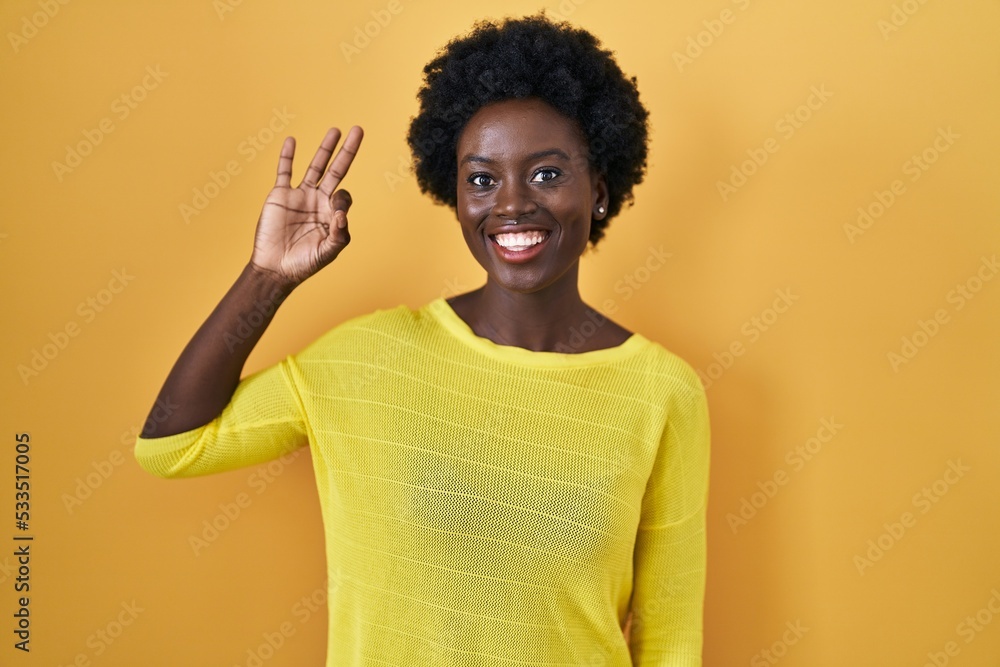 African young woman standing over yellow studio smiling positive doing ok sign with hand and fingers. successful expression. African young woman standing over yellow studio smiling positive doing ok sign with hand and fingers. successful expression.