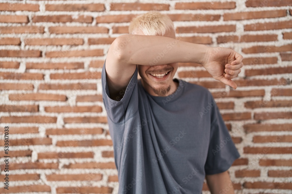 Young caucasian man standing over bricks wall covering eyes with arm smiling cheerful and funny. blind concept. Young caucasian man standing over bricks wall covering eyes with arm smiling cheerful and funny. blind concept.
