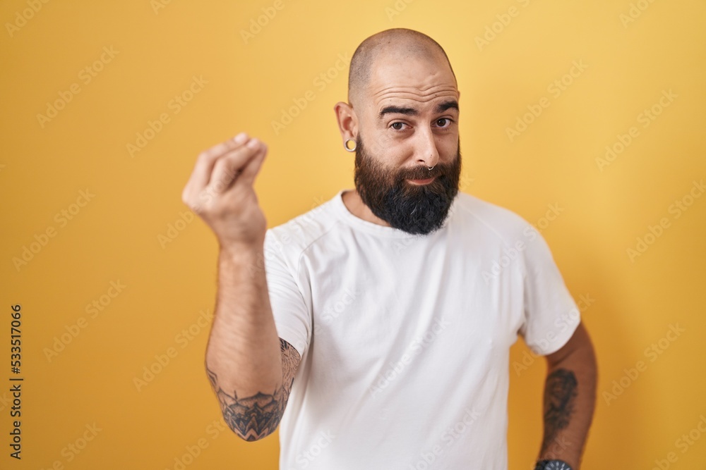 Young hispanic man with beard and tattoos standing over yellow background doing italian gesture with hand and fingers confident expression Young hispanic man with beard and tattoos standing over yellow background doing italian gesture with hand and fingers confident expression