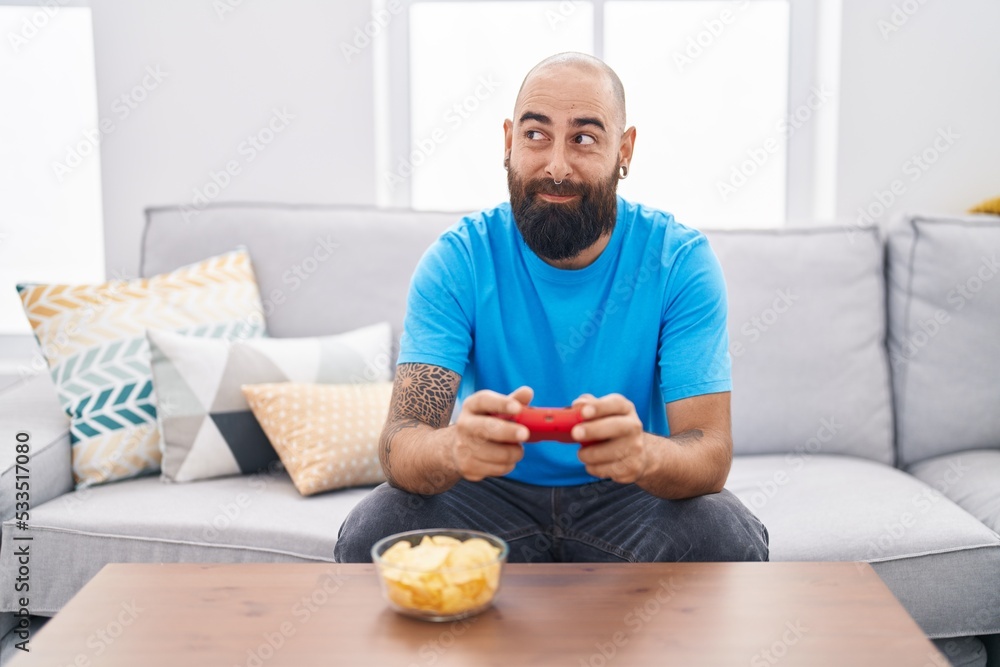 Young hispanic man with beard and tattoos playing video game sitting on the sofa smiling looking to the side and staring away thinking. Young hispanic man with beard and tattoos playing video game sitting on the sofa smiling looking to the side and staring away thinking.