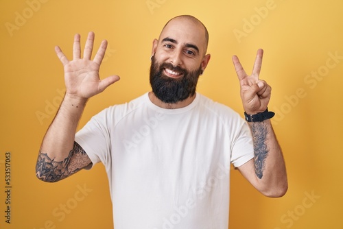 Young hispanic man with beard and tattoos standing over yellow background showing and pointing up with fingers number seven while smiling confident and happy.