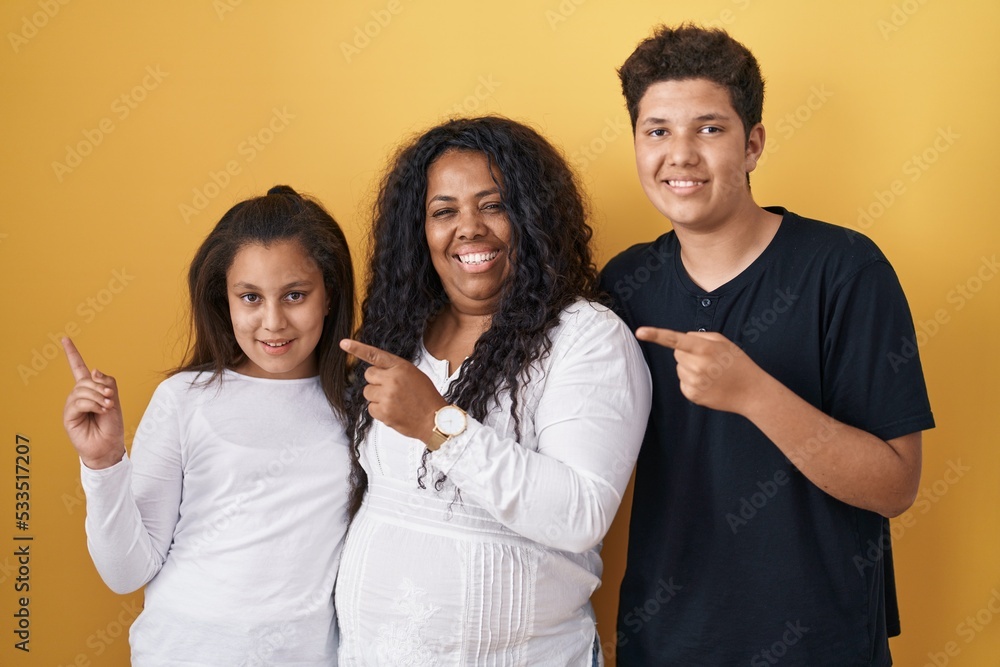 Family of mother, daughter and son standing over yellow background cheerful with a smile on face pointing with hand and finger up to the side with happy and natural expression Family of mother, daughter and son standing over yellow background cheerful with a smile on face pointing with hand and finger up to the side with happy and natural expression