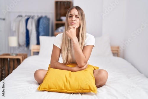 Young blonde woman sitting on the bed with pillow at home looking confident at the camera with smile with crossed arms and hand raised on chin. thinking positive.