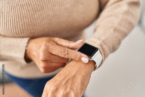 Middle age hispanic woman using smartwatch standing at home