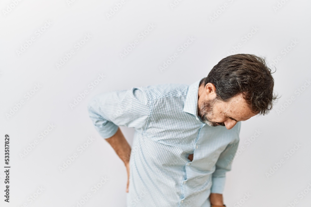 Middle age hispanic man with beard standing over isolated background suffering of backache, touching back with hand, muscular pain Middle age hispanic man with beard standing over isolated background suffering of backache, touching back with hand, muscular pain