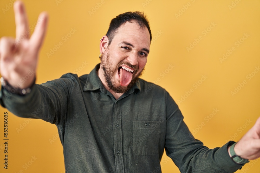 Plus size hispanic man with beard standing over yellow background smiling with tongue out showing fingers of both hands doing victory sign. number two. Plus size hispanic man with beard standing over yellow background smiling with tongue out showing fingers of both hands doing victory sign. number two.