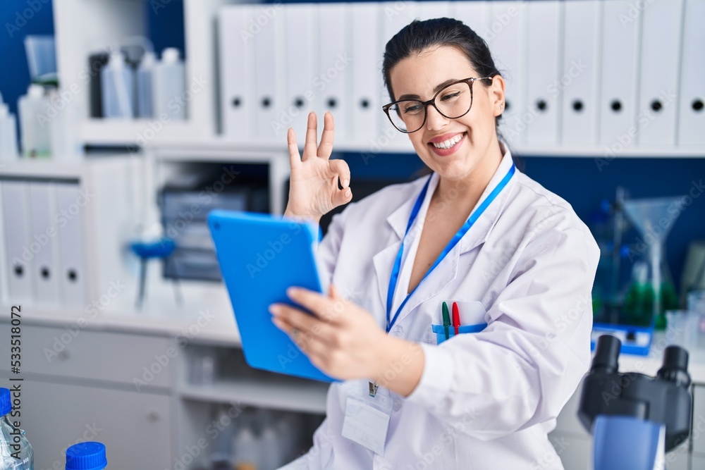 Young brunette woman working at scientist laboratory using tablet doing ok sign with fingers, smiling friendly gesturing excellent symbol Young brunette woman working at scientist laboratory using tablet doing ok sign with fingers, smiling friendly gesturing excellent symbol