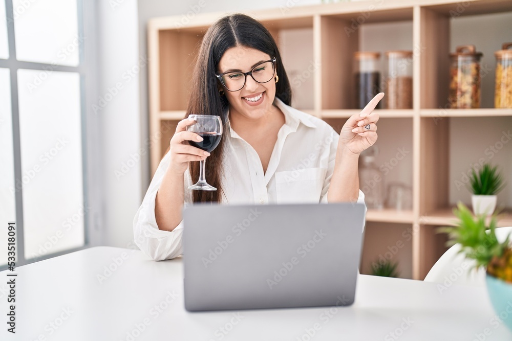 Young brunette woman doing video call with laptop drinking a glass of wine smiling happy pointing with hand and finger to the side Young brunette woman doing video call with laptop drinking a glass of wine smiling happy pointing with hand and finger to the side