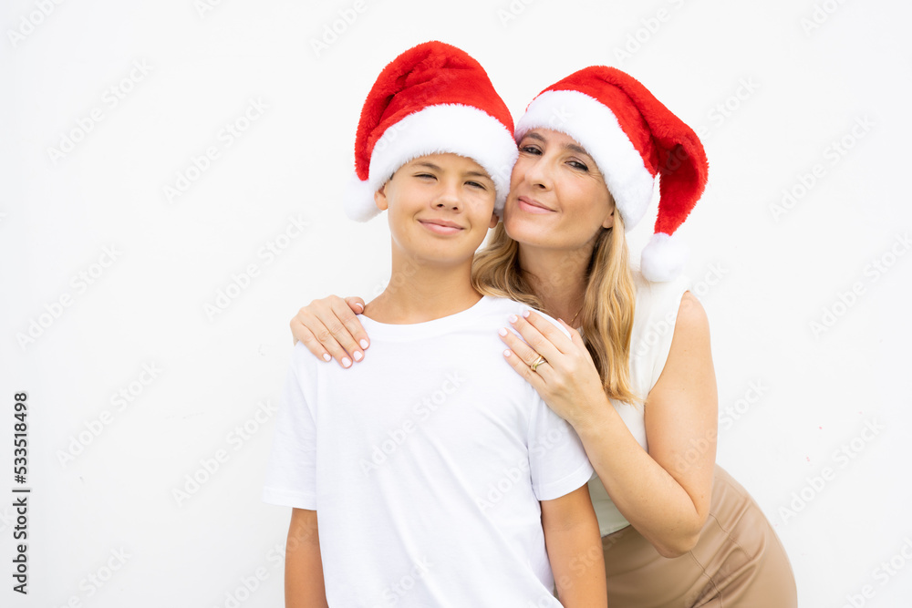 Obraz premium Pretty young mom and her handsome child son together wearing Santa hats against white wall outdoor