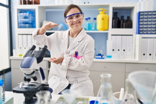Hispanic girl with down syndrome working at scientist laboratory gesturing with hands showing big and large size sign, measure symbol. smiling looking at the camera. measuring concept.
