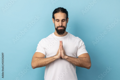Man with beard wearing white T-shirt concentrating his mind, keeping hands namaste gesture, meditating, yoga exercise breath technique reduce stress. Indoor studio shot isolated on blue background.