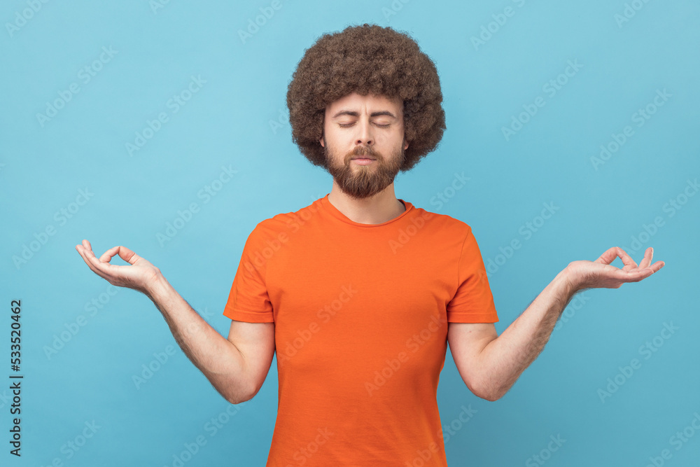Portrait of calm meditative handsome man with Afro hairstyle wearing orange T-shirt standing with closed eyes and yoga gesture pose. Indoor studio shot isolated on blue background. Portrait of calm meditative handsome man with Afro hairstyle wearing orange T-shirt standing with closed eyes and yoga gesture pose. Indoor studio shot isolated on blue background.