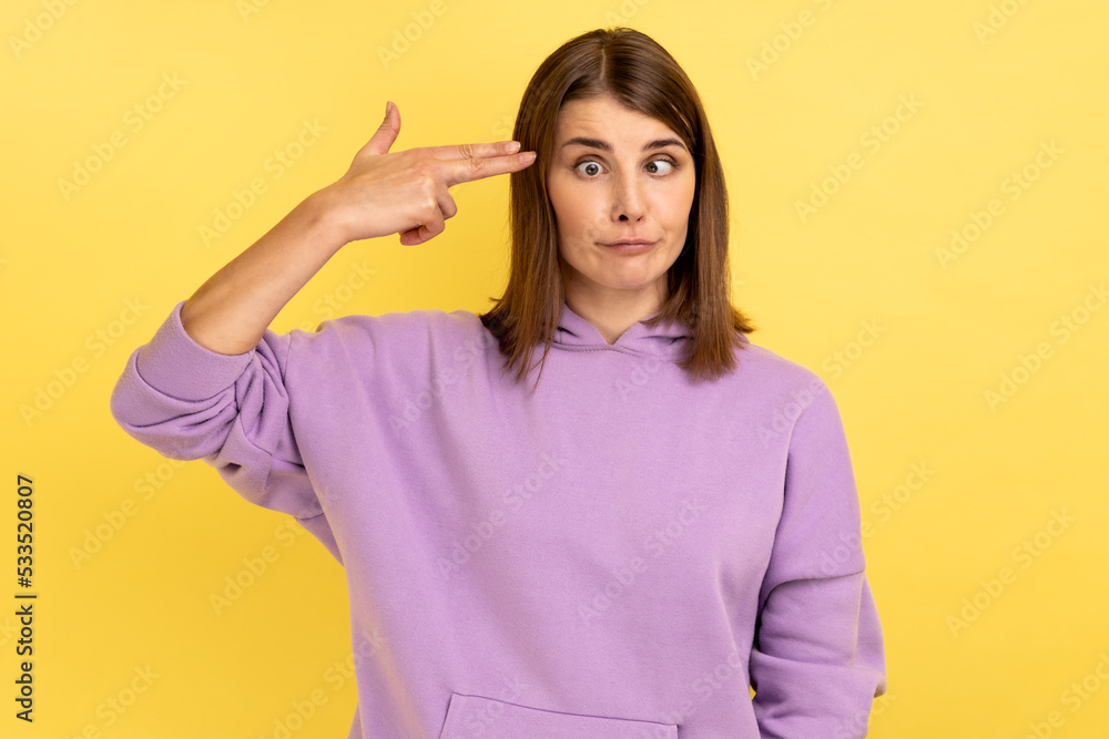Portrait of desperate woman, pointing finger gun to head, shooting herself, making suicide gesture to stop depression, wearing purple hoodie. Indoor studio shot isolated on yellow background. Portrait of desperate woman, pointing finger gun to head, shooting herself, making suicide gesture to stop depression, wearing purple hoodie. Indoor studio shot isolated on yellow background.