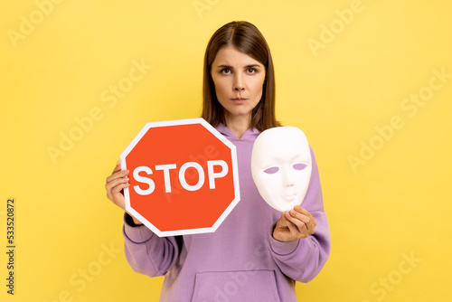 Portrait of serious woman holding red stop sign and white mask, looking at camera with strict bossy expression, wearing purple hoodie. Indoor studio shot isolated on yellow background. photo