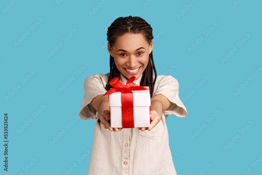 Portrait of happy woman with black dreadlocks giving present box, looking at camera, congratulating friend with birthday, wearing white shirt. Indoor studio shot isolated on blue background. Portrait of happy woman with black dreadlocks giving present box, looking at camera, congratulating friend with birthday, wearing white shirt. Indoor studio shot isolated on blue background.