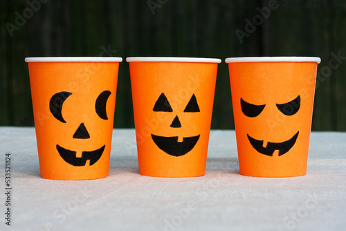 Front view of Halloween crafts, three orange paper cups for drinks with painted faces stand on table against background of wooden wall. Decor for Halloween.