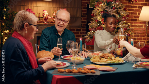 Happy elderly man talking with wife while enjoying Christmas dinner table with close family members. Multiethnic festive people celebrating traditional winter holiday while eating home cooked food.