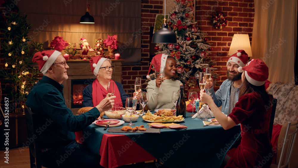 Happy family members wearing festive hats while clinking glasses with champagne. Joyful diverse people laughing heartily while enjoying Christmas dinner together at home. Happy family members wearing festive hats while clinking glasses with champagne. Joyful diverse people laughing heartily while enjoying Christmas dinner together at home.