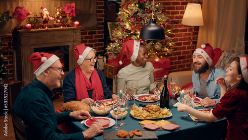 Cheerful laughing heartily multiethnic family toasting sparkling wine while singing Christmas songs at dinner. Festive diverse people wearing traditional hats enjoying winter holiday together at home.
