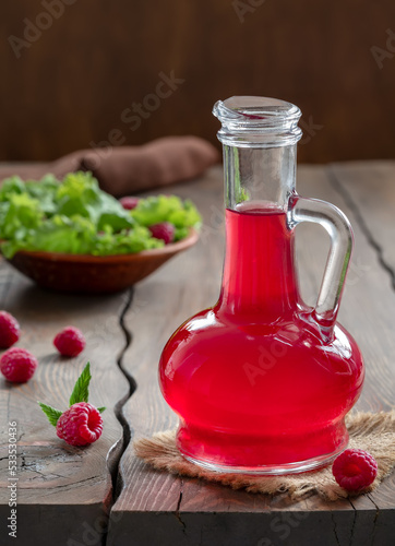 Glass cruet of raspberry vinegar or vinaigrette salad dressing on wooden table with fresh berries and plate of salad on background. Vertical, selective focus. photo
