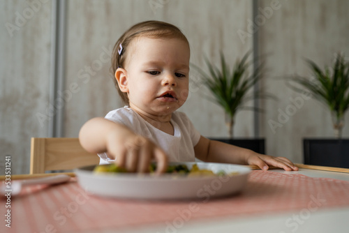 One baby girl small caucasian toddler eating while sit at the table alone at home copy space little child having lunch healthy food meal in room in day serious concentrated