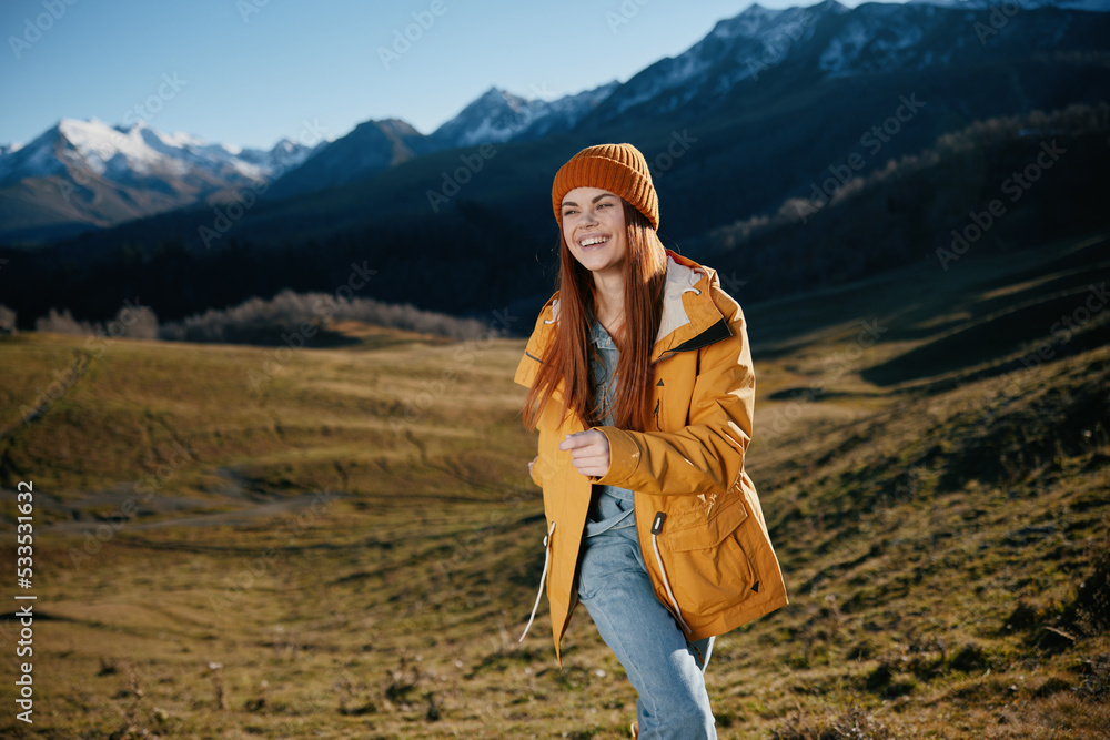 Woman beautifully running up the hill to the camera smile with teeth in the mountains in the autumn in a yellow raincoat and jeans happy sunset trip on a hike mountains in the snow, freedom lifestyle