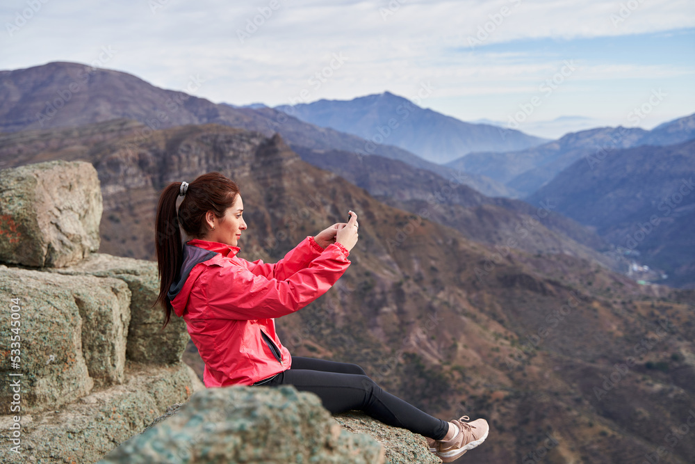 young woman sideways taking a picture with her smartphone, wearing a red jacket, sitting on a rock in the middle of the Andes mountain range in Chile