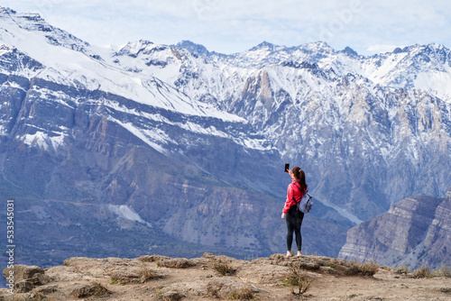 young red haired woman with red jacket and backpack, taking pictures with her phone in the middle of the Andes mountain range in Chile © daniel