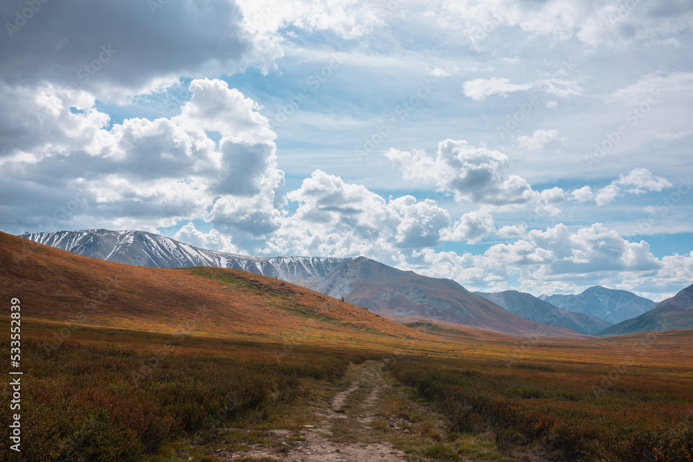 Motley autumn landscape with hills on high plateau and sunlit snowy mountain range under dramatic cloudy sky. Vivid autumn colors in mountains. Sunlight and shadows of clouds in changeable weather.