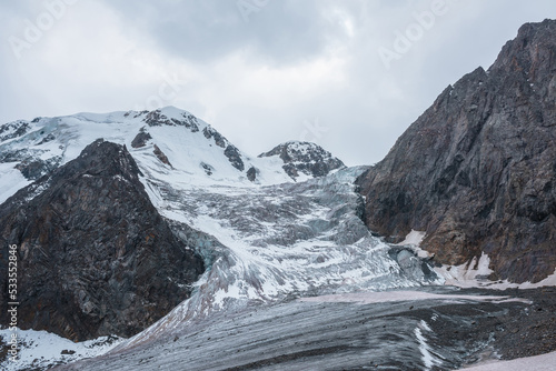 Dramatic landscape with glacier icefall on large snow mountain range with sharp rocky pinnacle under gray cloudy sky. Big glacier close-up in high altitude. Gloomy scenery in mountains in overcast.
